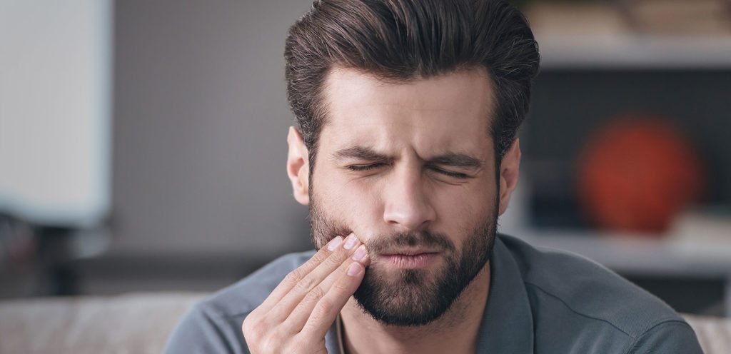 Frustrated young man touching his cheek and keeping eyes closed while sitting on the couch at home